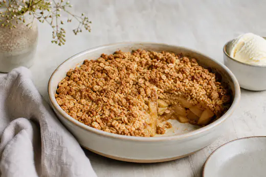 Goldbrauner Apple Crumble in einer runden Auflaufform, ein Stück ausgestochen, mit Vanilleeis daneben